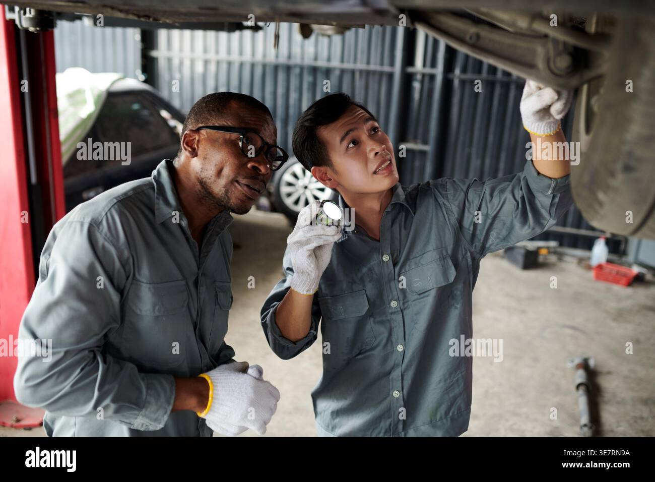 Meccanici maschi neri e asiatici che indossano guanti che riparano sotto il telaio dell'auto mentre si trova in un'officina di riparazione auto Foto Stock