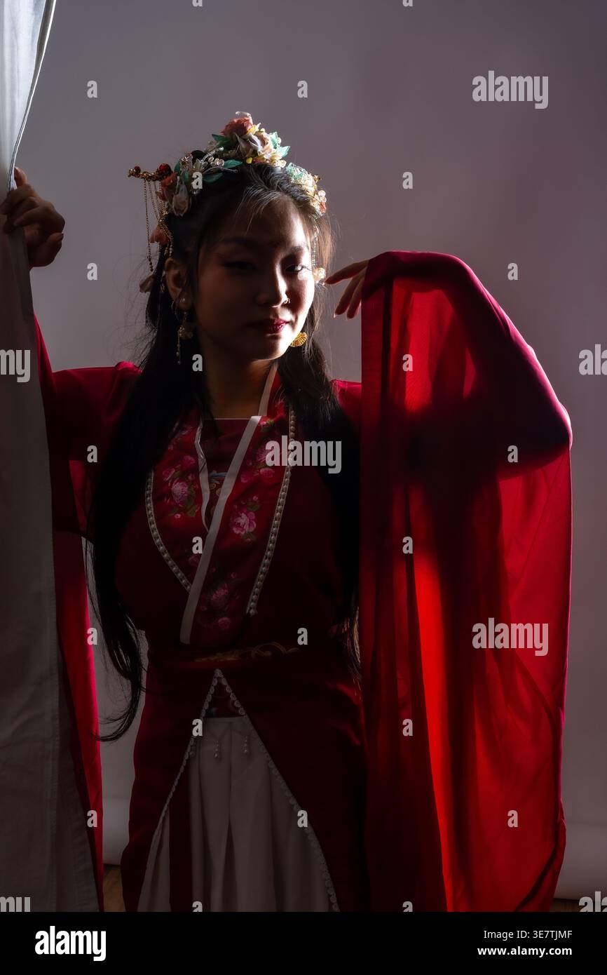 Donna asiatica in abito rosso tradizionale che indossa la corona di fiori tirando la tenda da da parte mentre si sta in piedi con la mano sollevata vicino al viso contro il taupe viola dietro... Foto Stock