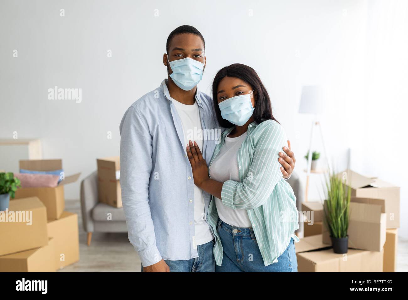 Coppia nera che indossa maschere protettive per il viso che si abbraccia mentre sta in piedi tra le scatole di cartone nel soggiorno di casa Foto Stock