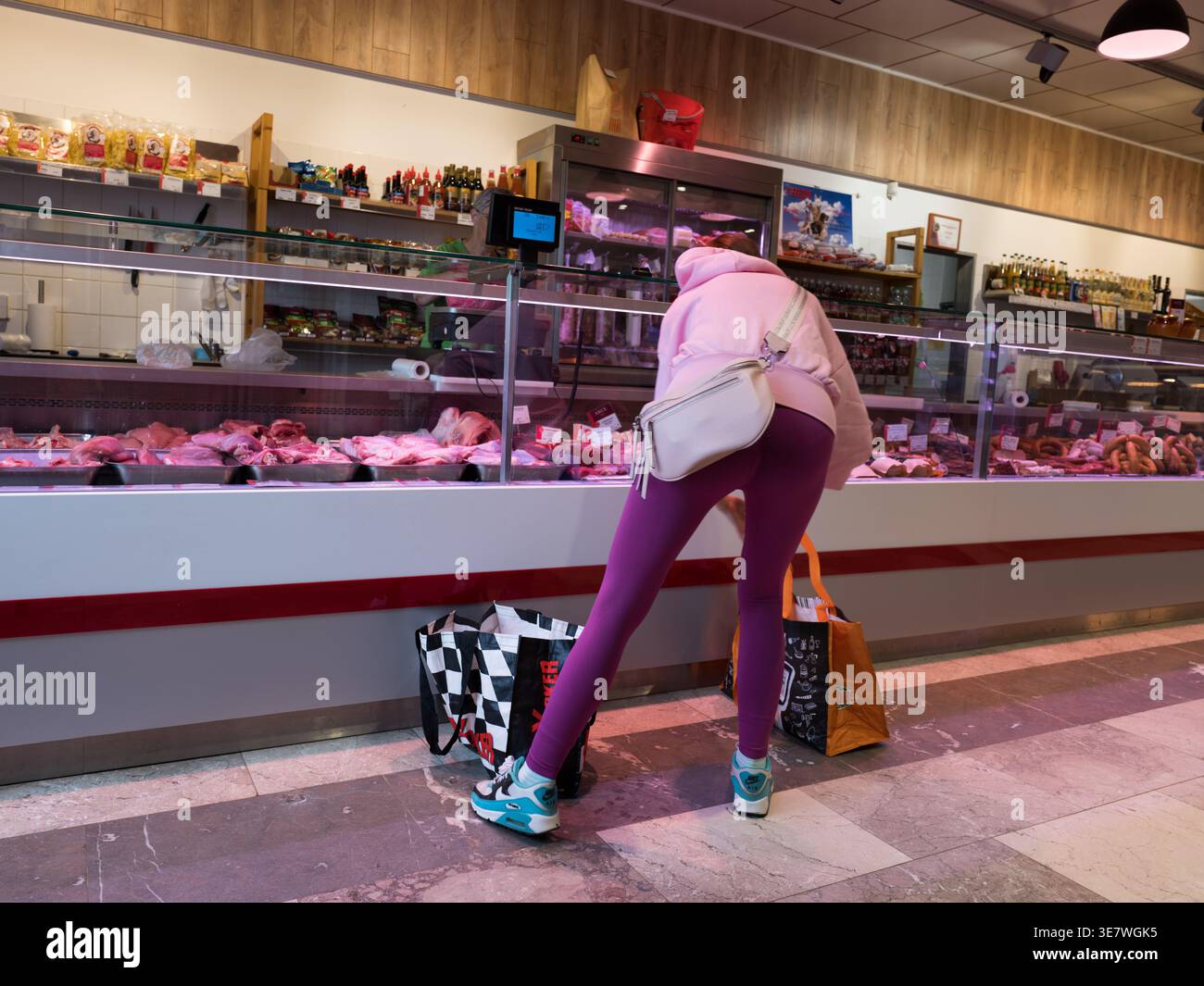 Donna con felpa con cappuccio rosa e leggings viola che guarda la carne fresca in un banco di macelleria durante lo shopping Foto Stock