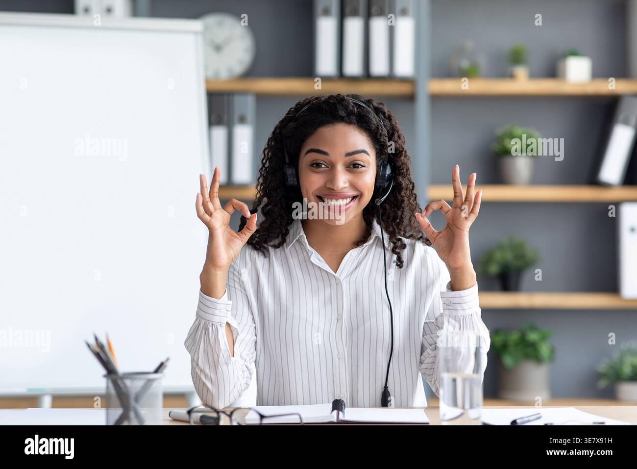 Donna nera con capelli ricci che indossano le cuffie che fanno un buon gesto della mano seduta al tavolo dell'ufficio domestico Foto Stock