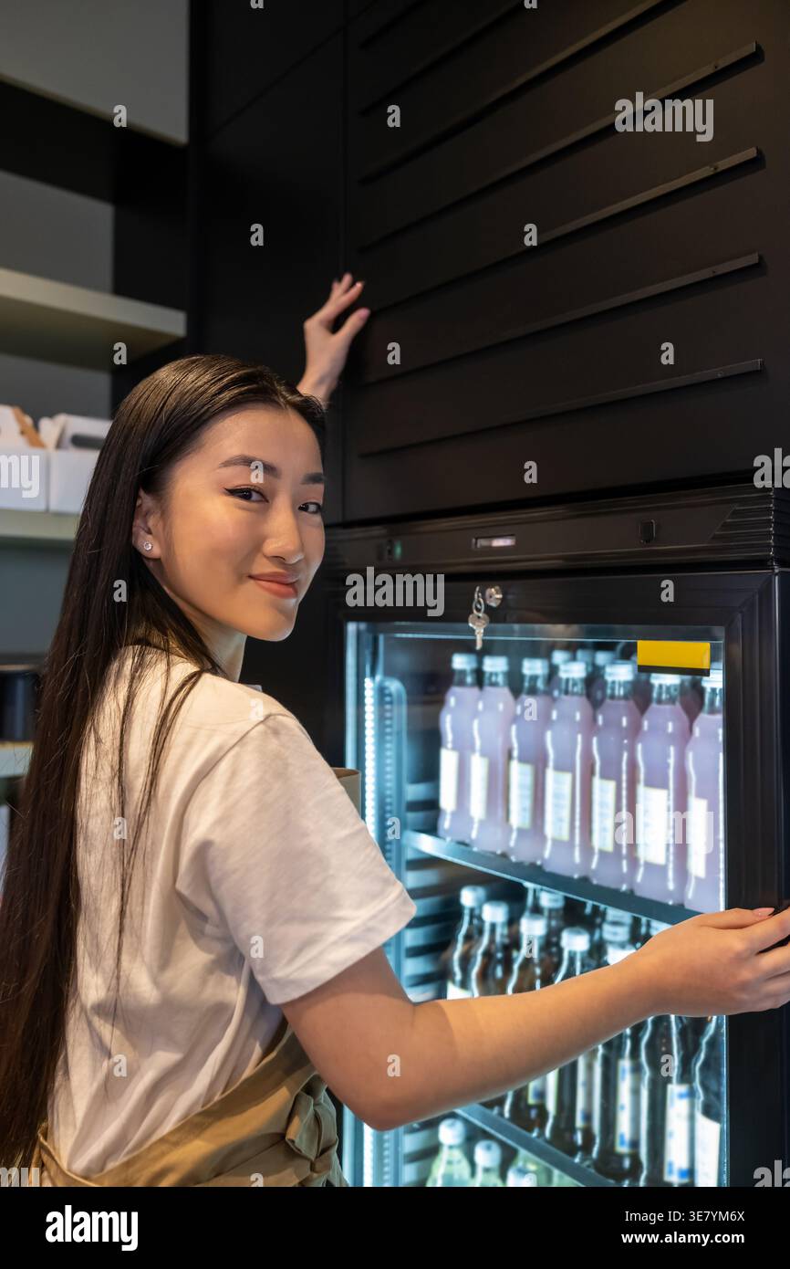 Sorridente lavoratrice asiatica del caffè in piedi sulla scala a cavalletto e toccando il muro sopra il mobile refrigerato con bevande analcoliche nel caffè Foto Stock