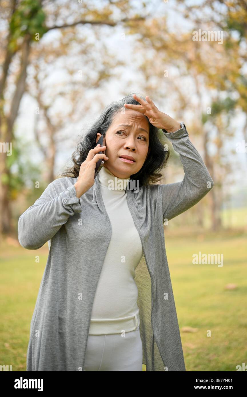 Donna asiatica anziana che guarda lontano toccando la fronte, mentre parla al telefono in piedi sul prato del parco durante il giorno Foto Stock