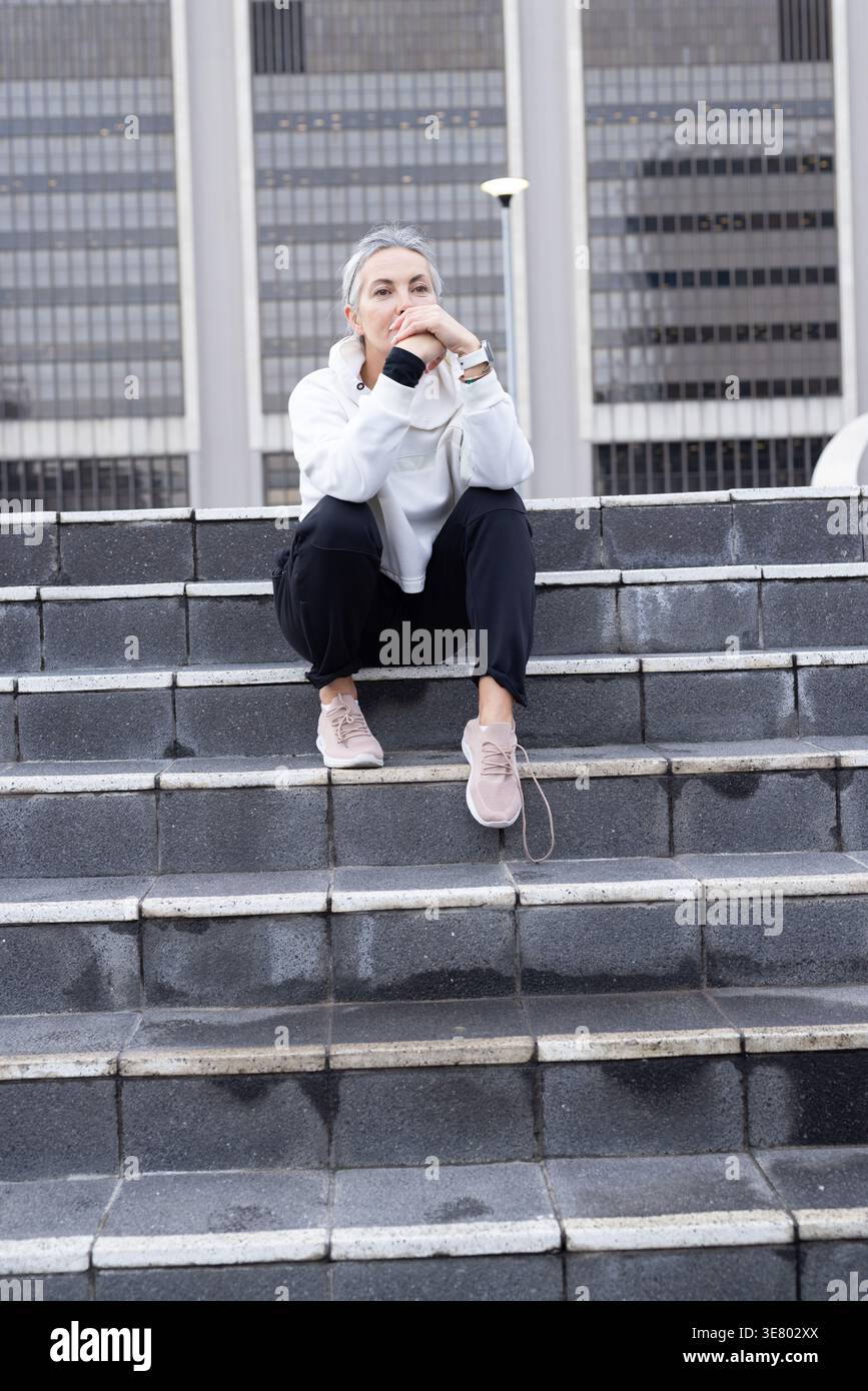 Donna anziana seduta su gradini di pietra nella piazza urbana, indossa felpa con cappuccio, riposa e guarda dopo la pioggia Foto Stock