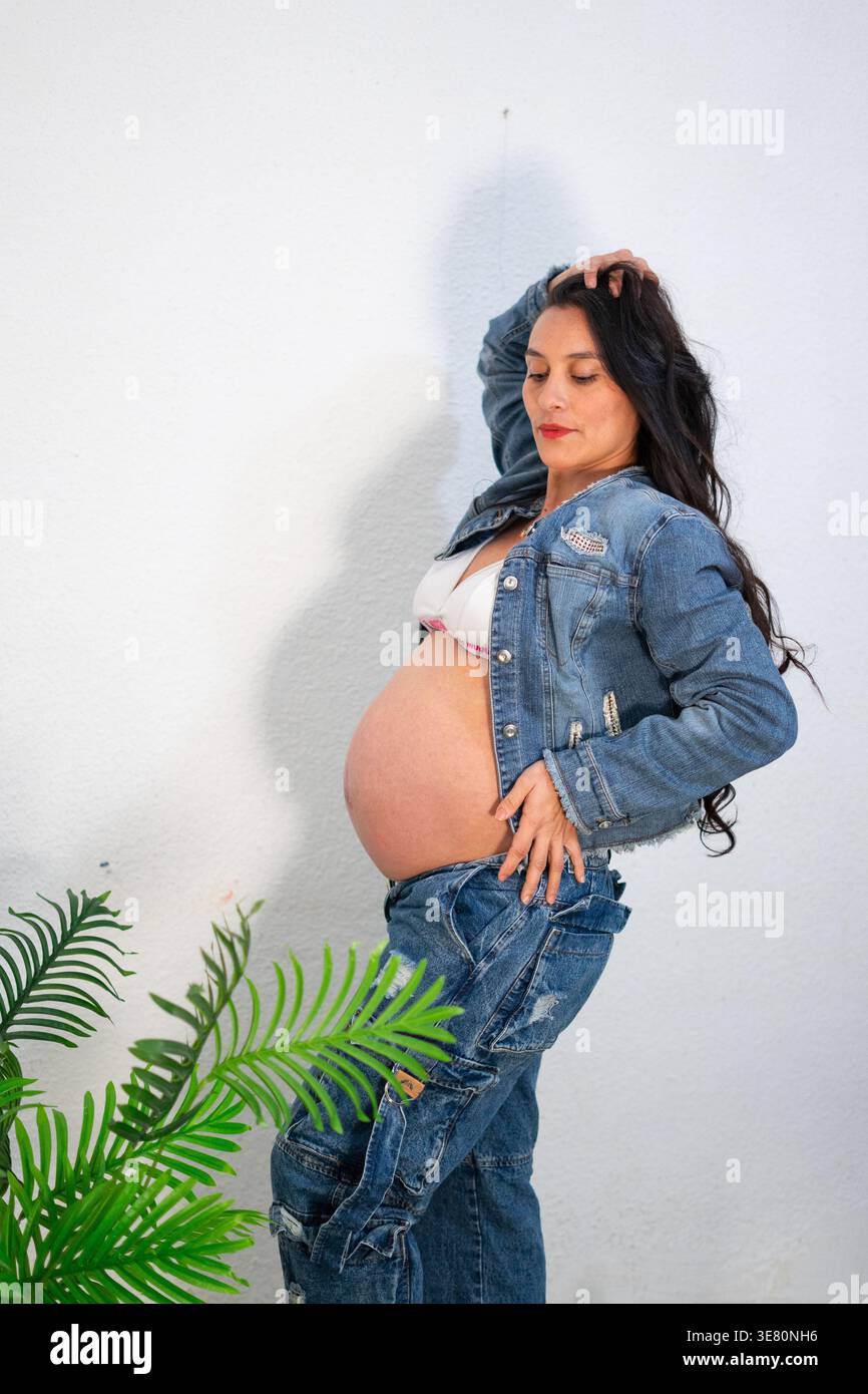 Donna incinta ispanica in piedi con la mano sui capelli e l'anca vicino alle piante in studio Foto Stock