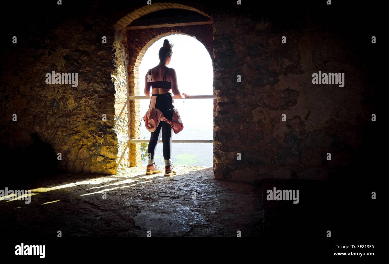 Donna in piedi all'interno di una rovina di pietra scura, che guarda il luminoso paesaggio all'aperto attraverso una finestra ad arco. Contemplazione e viaggio. Vista esplorativa Foto Stock