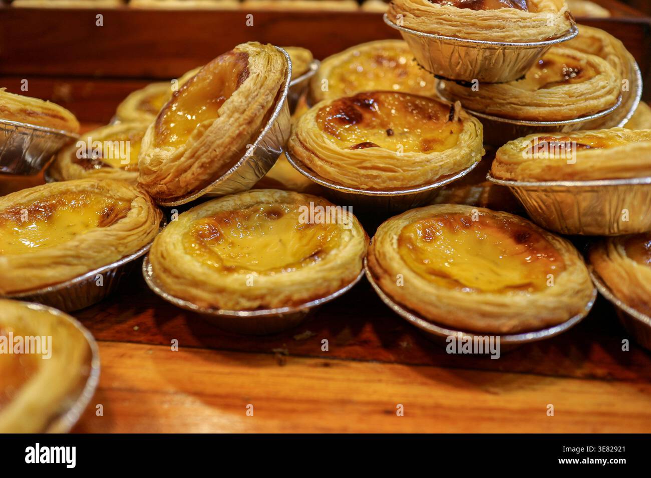 Pila di crostate di uova portoghesi appena sfornate in tazze di alluminio su sfondo di legno. Crosta dorata friabile con crema pasticcera caramellata per prodotti da forno e alimenti. Foto Stock