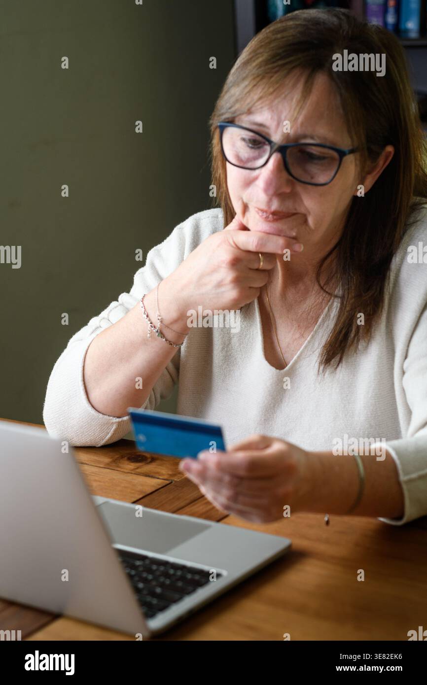 Donna anziana che pensa mentre tiene in mano la carta di credito e utilizza il laptop, prendendo in considerazione la decisione di acquisto online. Foto Stock