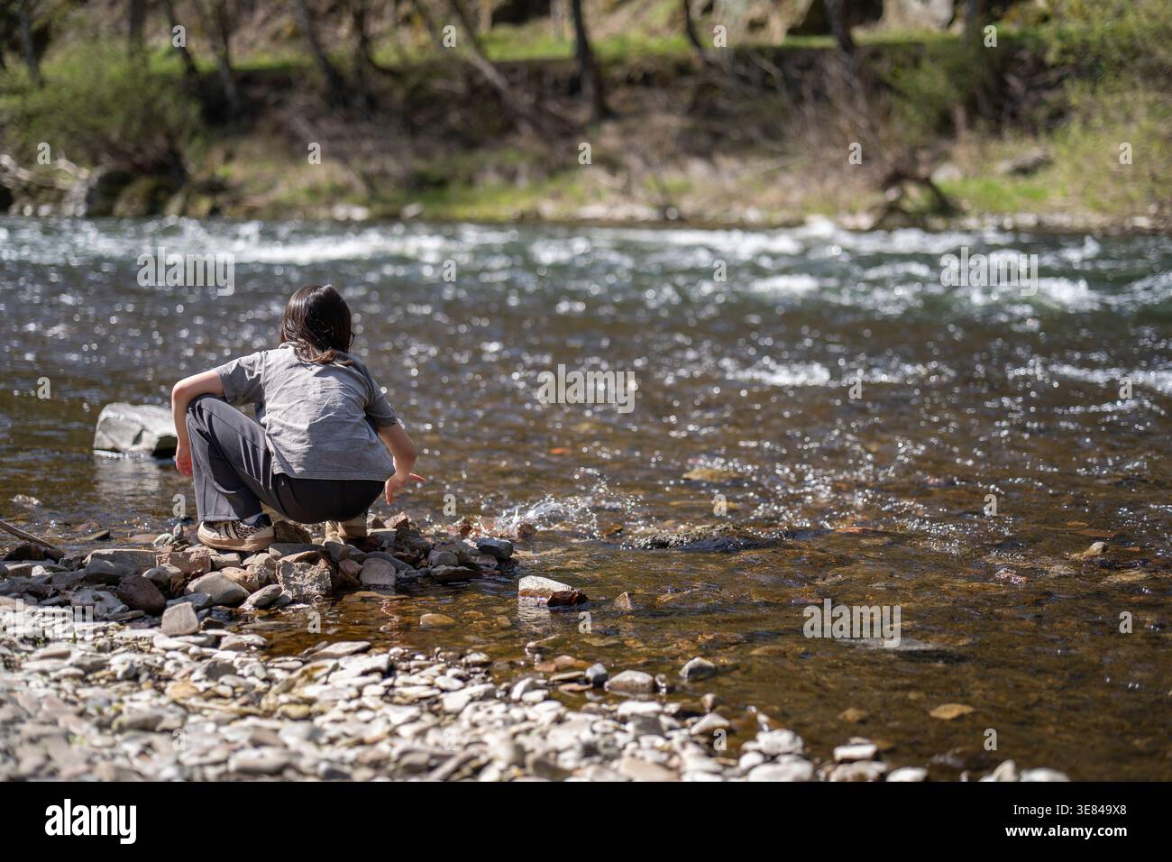 Bambino accovacciato sulla riva rocciosa del fiume che tocca l'acqua. Concetto di apprendimento tattile Foto Stock