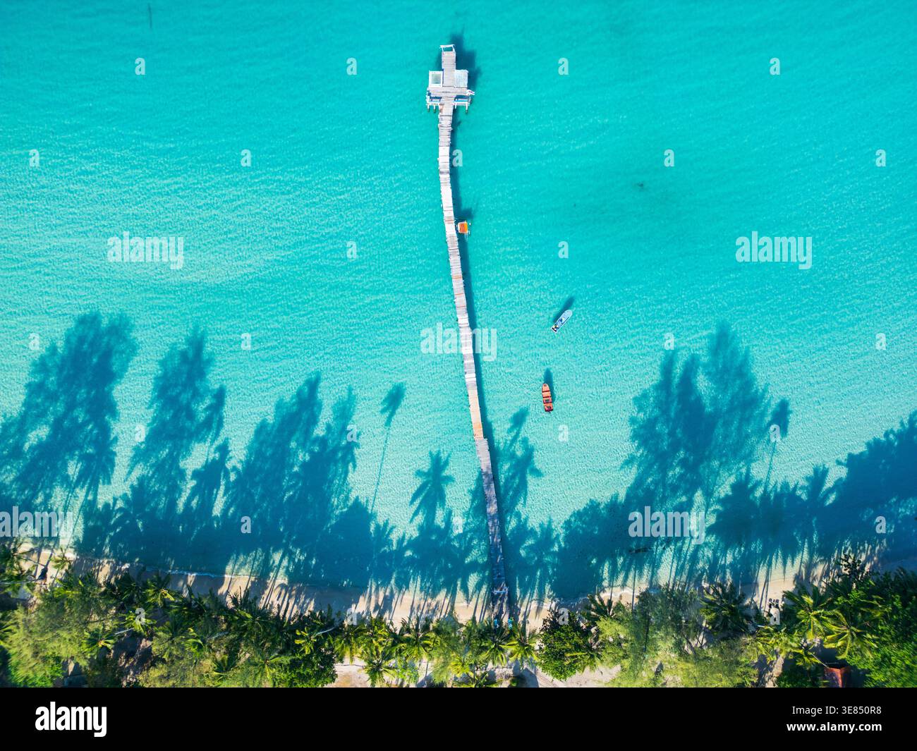 Vista aerea dall'alto verso il basso di un lungo molo che si estende nel mare turchese con piccole barche nelle vicinanze e ombre di palme lungo una costa tropicale sabbiosa. Foto Stock