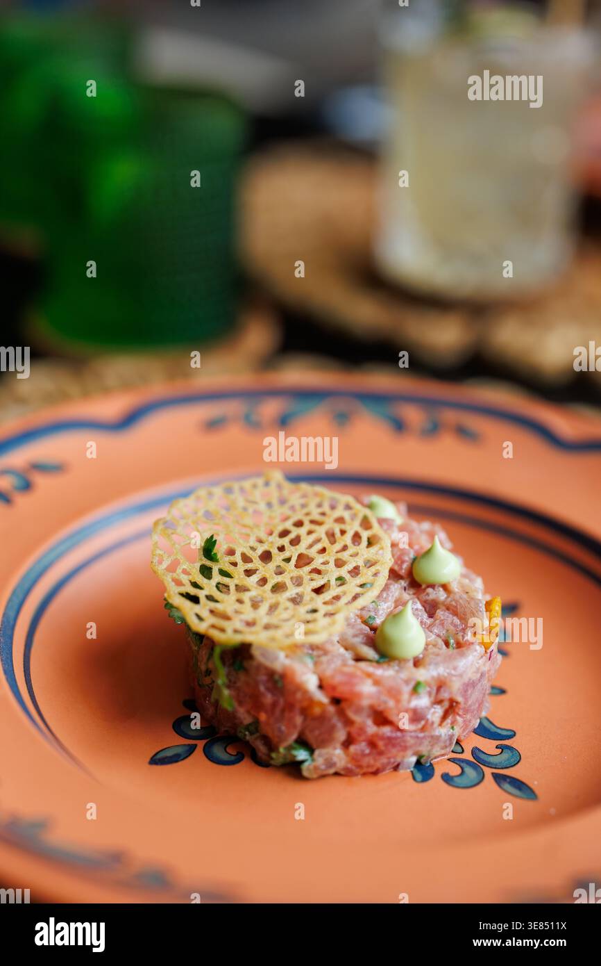 Tartare di bistecca da buongustai placcato su un piatto decorativo in terracotta con punti di salsa e decorazioni croccanti in tuile; profondità di campo bassa con bevande sfocate sul retro Foto Stock