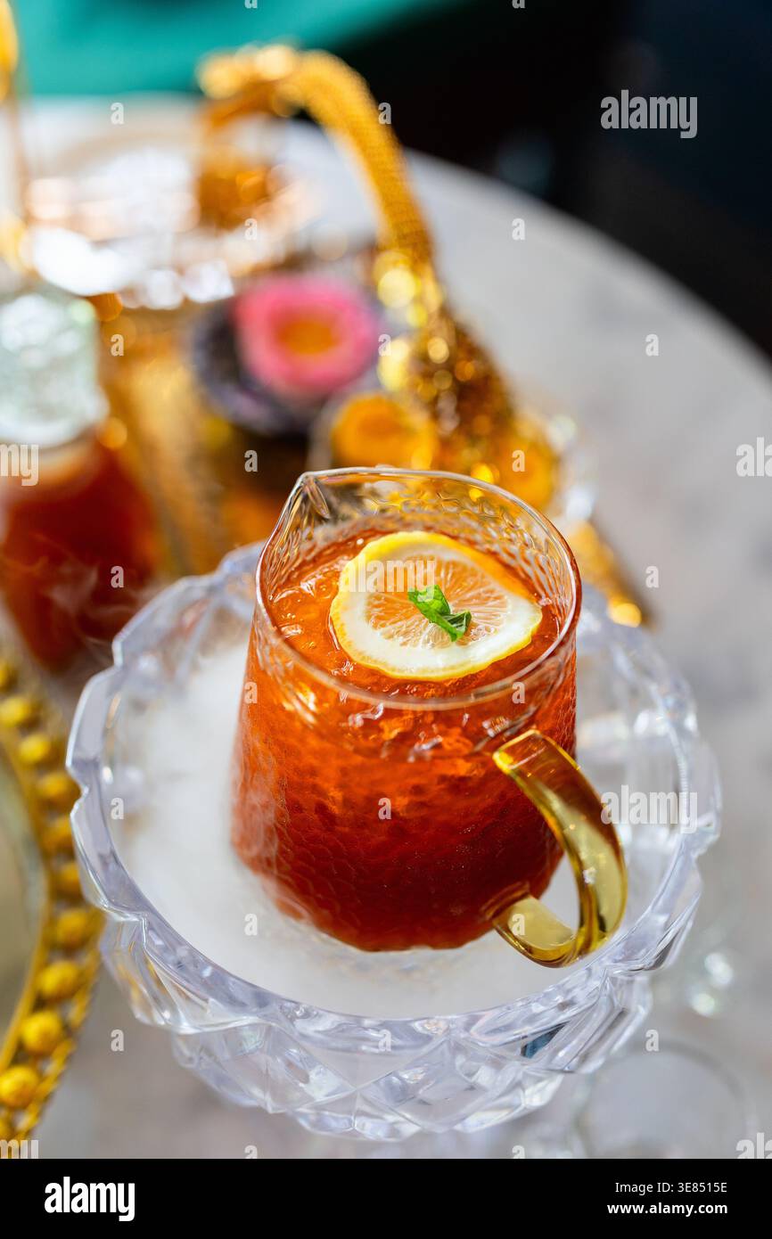 Tazza di tè freddo in vetro con fetta di limone e menta su piattino di cristallo, manico dorato, sul tavolo da caffè con vassoio teiera decorato bokeh. Foto Stock