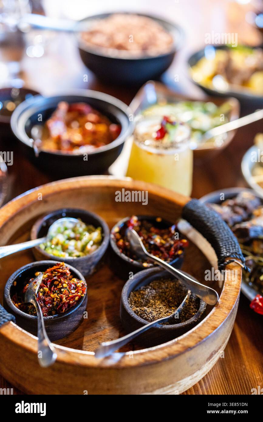 Primo piano di spezie assortite e condimenti al peperoncino in piccole ciotole nere su un vassoio di legno al tavolo del ristorante con piatti sfocati e bevande all'interno Foto Stock