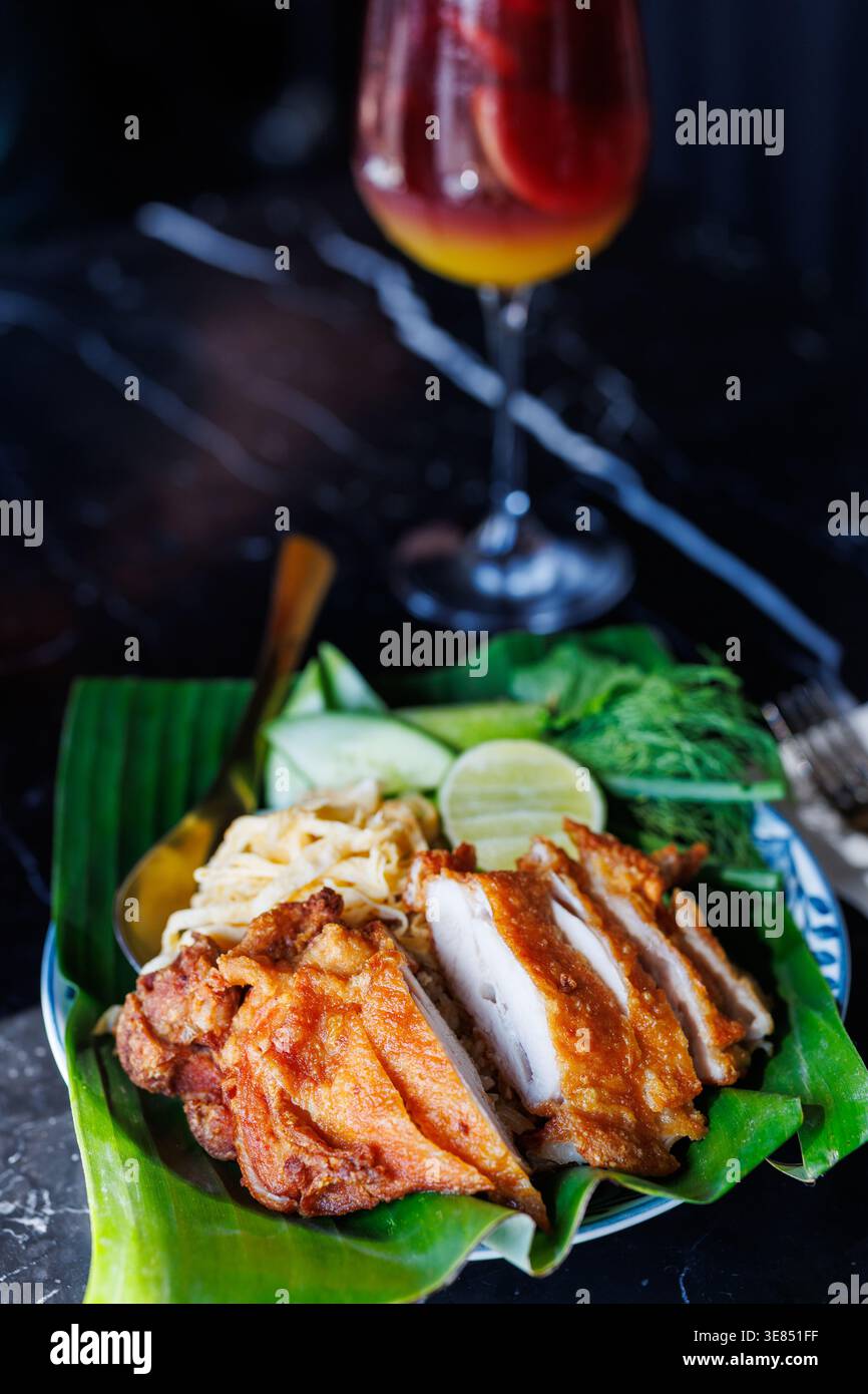 Pollo fritto tailandese servito su una foglia di banana con cetriolo, cavolo, lime ed erbe; cocktail colorato sullo sfondo su un tavolo di marmo scuro, profondità bassa Foto Stock