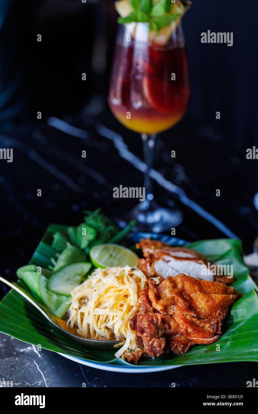 Pollo fritto in stile tailandese e insalata di papaya grattugiata su un piatto di foglie di banana con cetriolo, lime ed erbe aromatiche, serviti con un cocktail rosso a strati in uno stelo Foto Stock