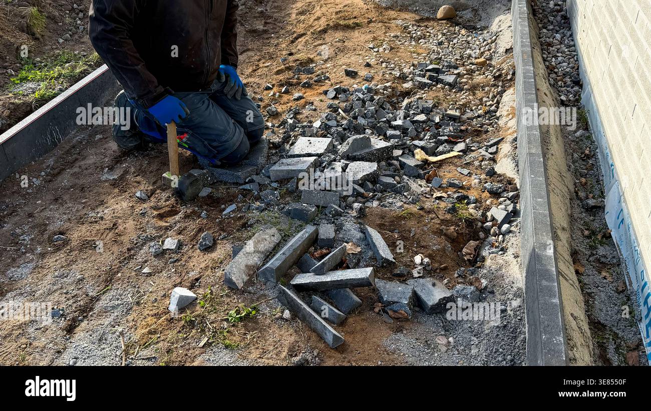 Lavoratore maschio inginocchiato sul terreno in prossimità di pezzi di cemento rotti, utilizzando un martello per scomporre pietre in un'area di costruzione con ghiaia e sporcizia visibili Foto Stock