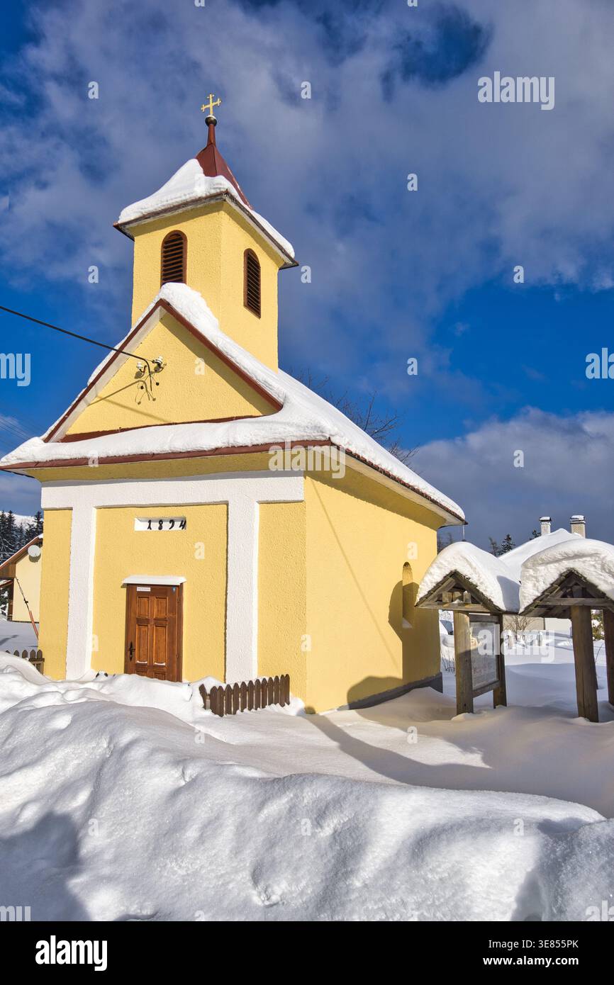 Veduta di una piccola e vibrante cappella gialla con un tetto rosso accoccolata nella neve contro un cielo blu brillante, Polianka, Banskobystricky kraj, Slovacchia. Foto Stock