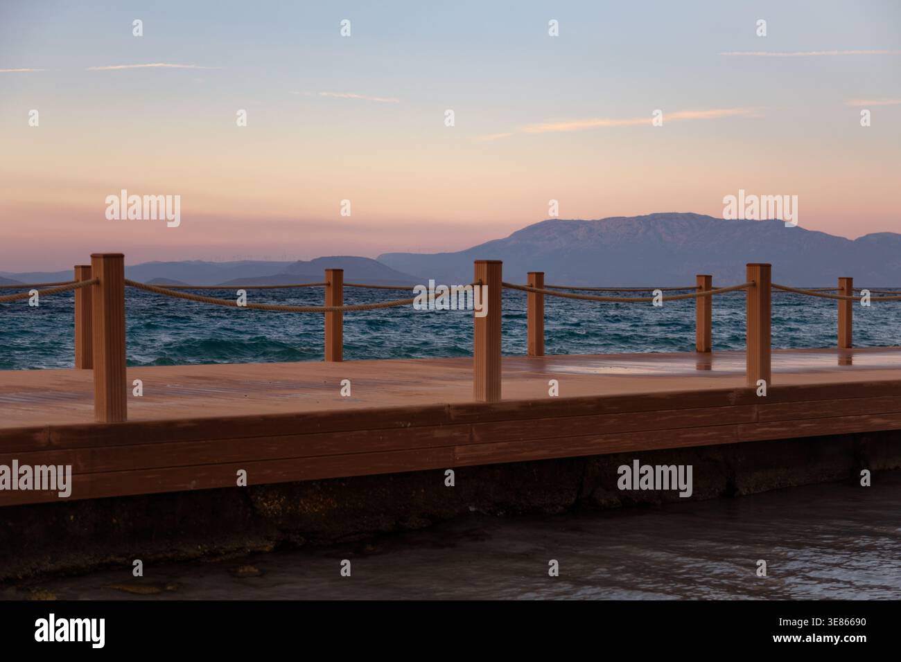 Un tranquillo molo sul mare si estende su dolci onde mentre il sole tramonta, gettando calde sfumature sull'acqua tranquilla e sulle montagne lontane. Una mamma pacifica Foto Stock
