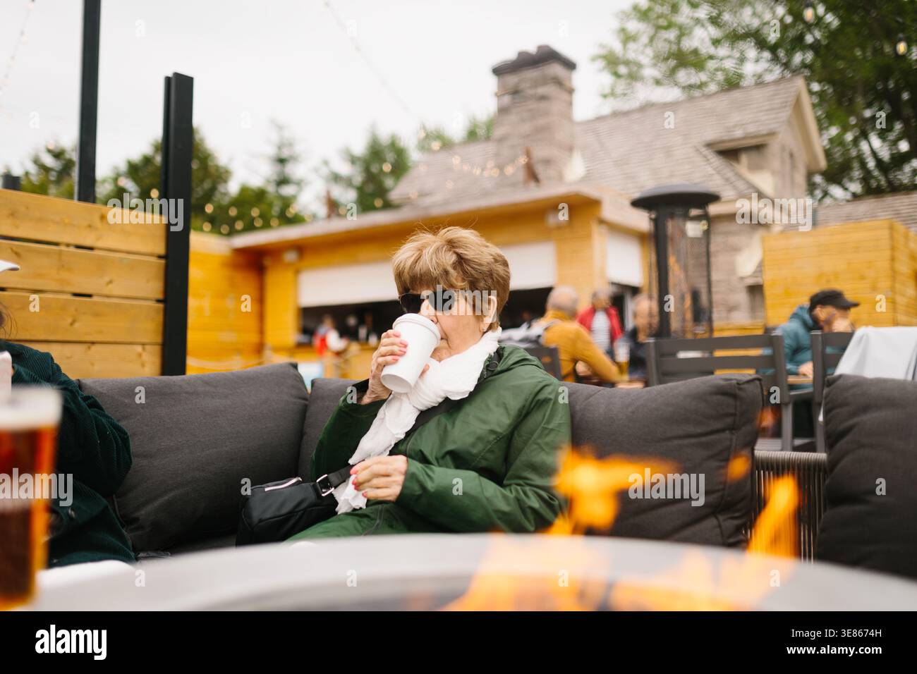 Una donna anziana con occhiali da sole e giacca verde beve caffè caldo in un accogliente patio all'aperto, seduto su un divano vicino a un caminetto con gli ospiti offuscati del caffè behi Foto Stock