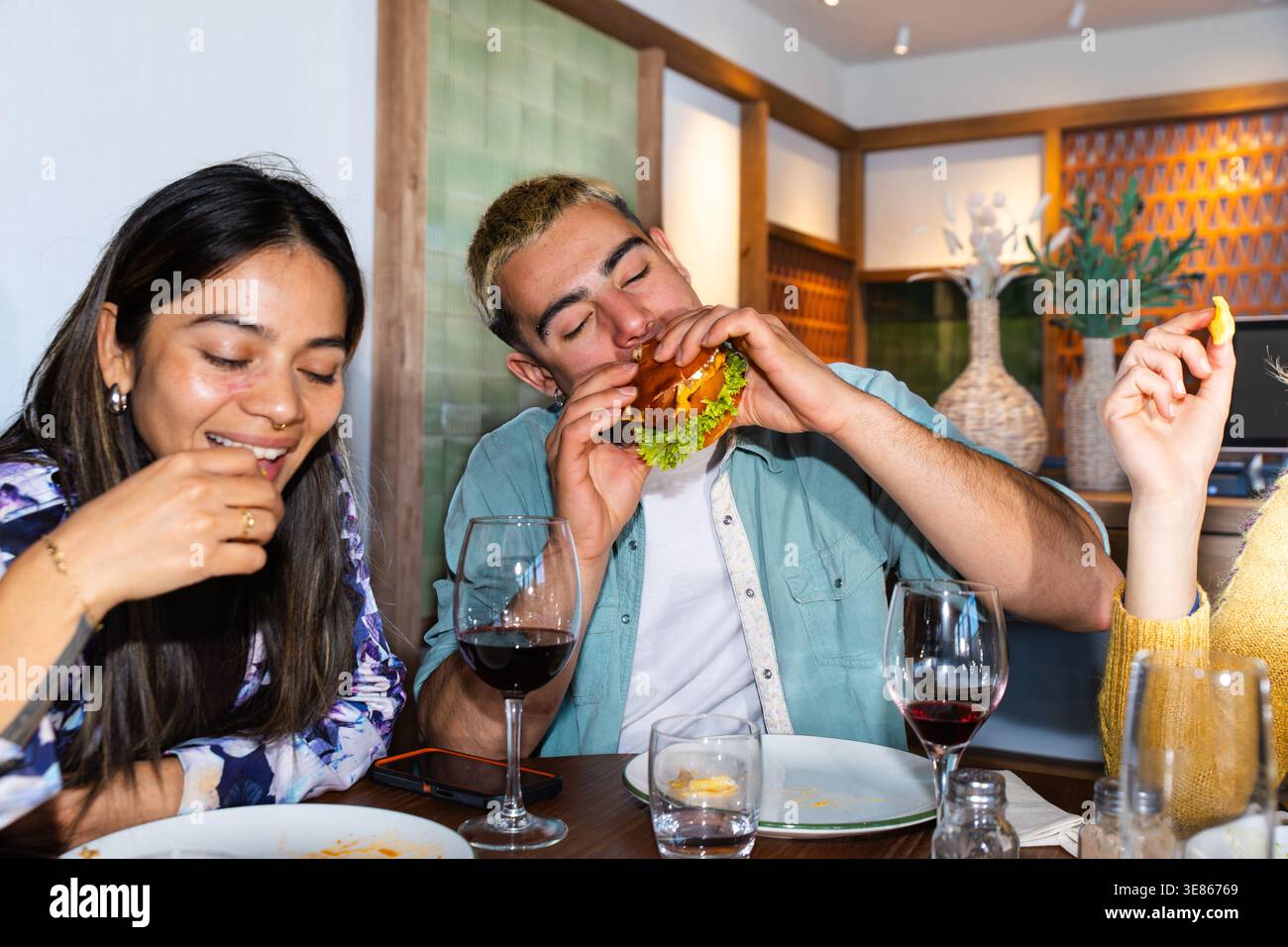 Gruppo di amici che si gusta una cena informale in un ristorante, condividendo hamburger, patatine fritte e vino. Momenti rilassanti di vita gastronomica con risate, conversati Foto Stock