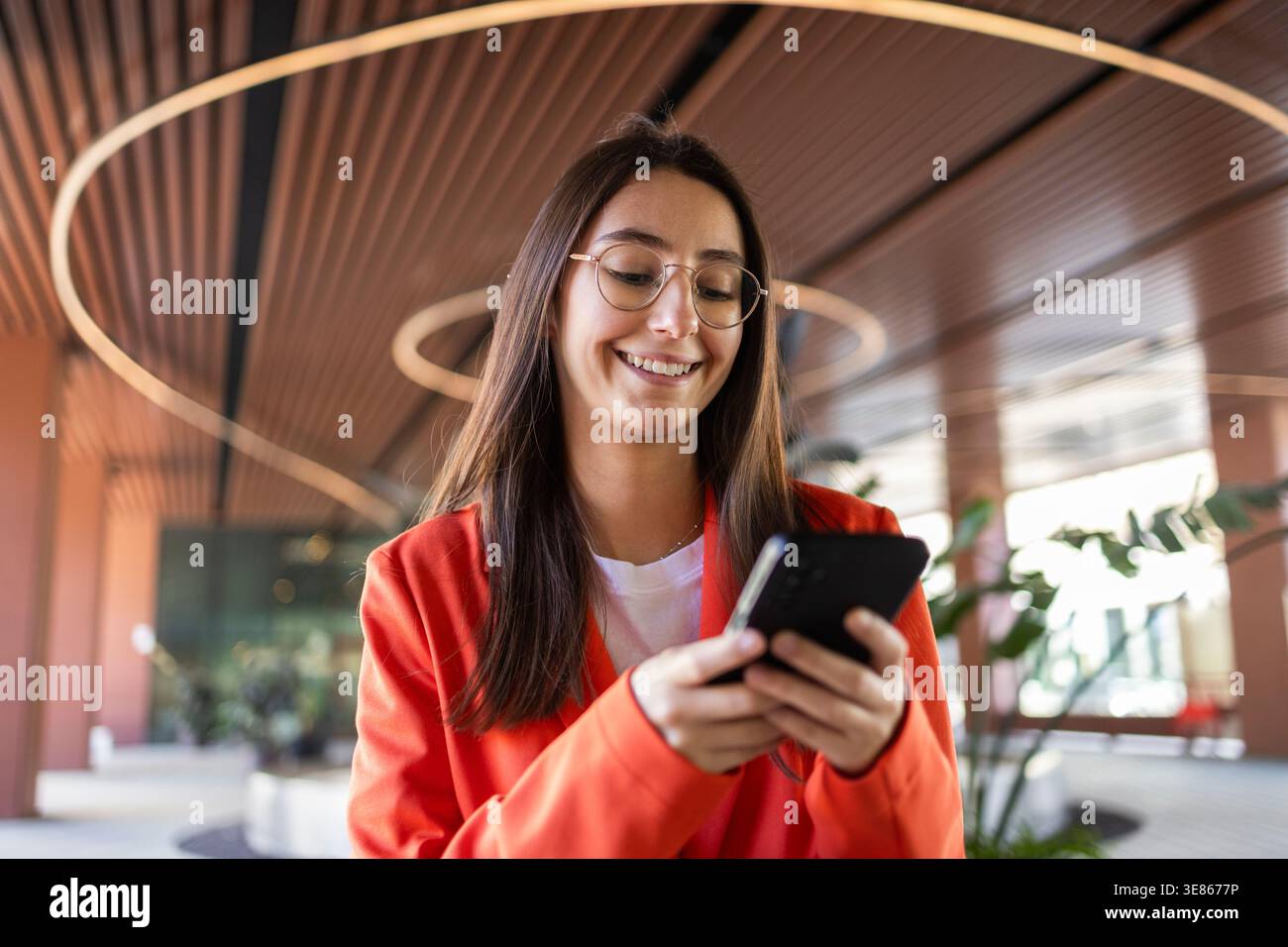 Una giovane donna d'affari con un blazer arancione sorride mentre usa il suo smartphone all'interno di un moderno edificio di uffici, mostrando un desiderio professionale e allegro Foto Stock