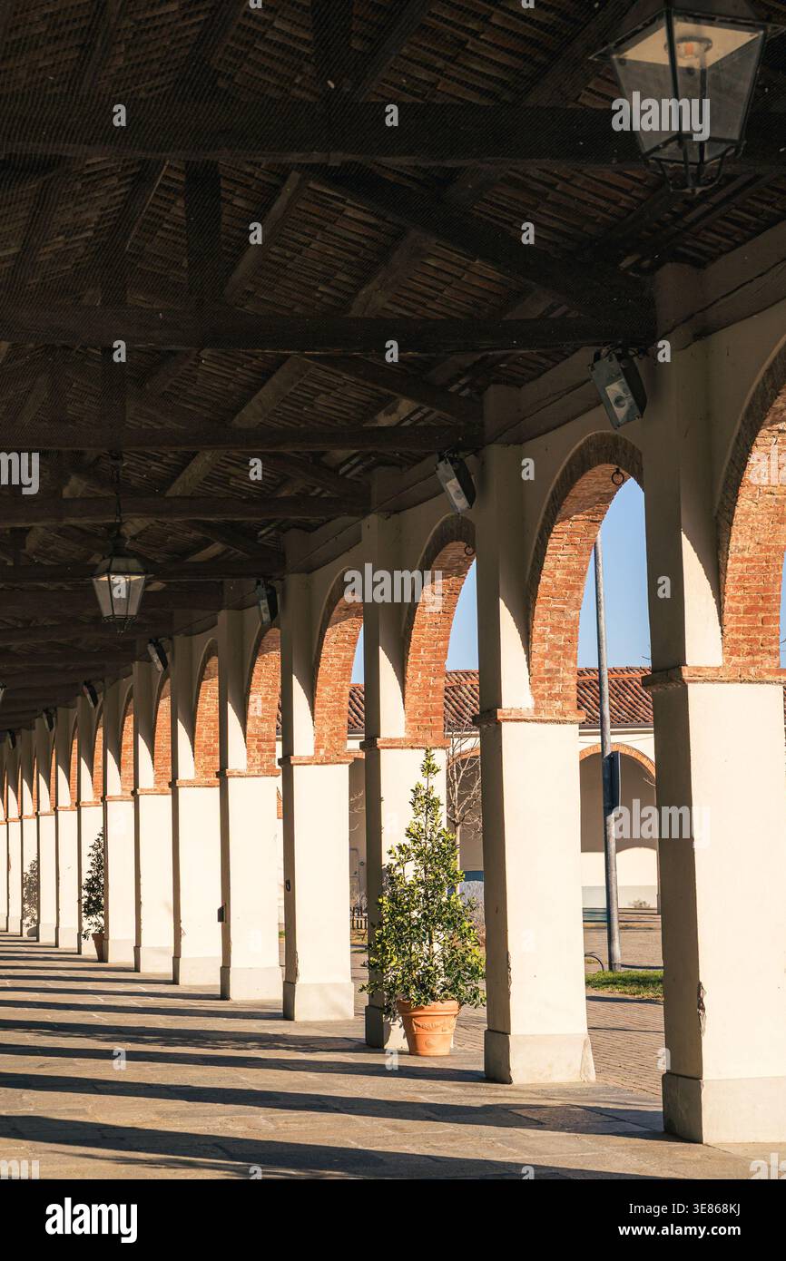 La luce del sole mattutina filtra attraverso gli archi di un classico colonnato italiano a Piamonte, Italia. La struttura in mattoni proietta ombre ripetute sul gr Foto Stock