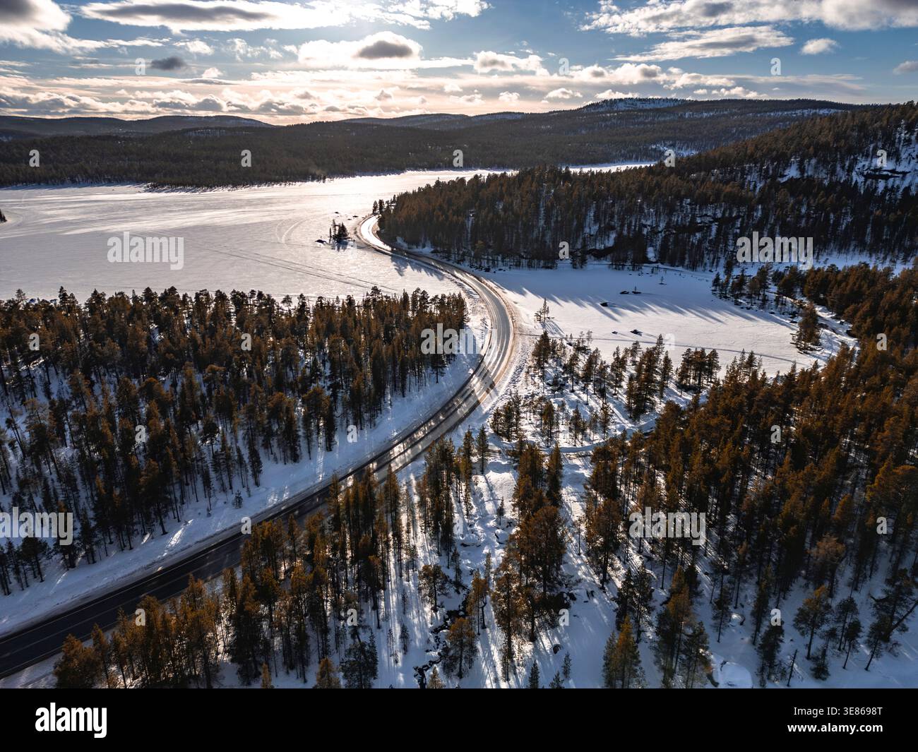 Vista su una strada tortuosa che attraversa la pineta innevata accanto a un lago ghiacciato in Finlandia, con basso sole invernale, lunghe ombre e nuvole spettacolari. Foto Stock