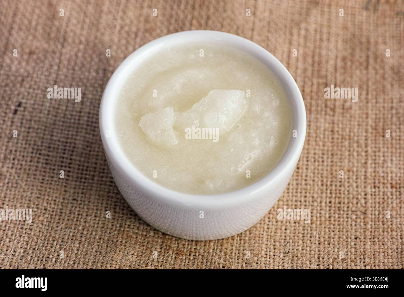 Una ciotola in ceramica bianca riempita con olio di cocco bianco puro posta su una superficie rustica in tessuto burlap. Primo piano. Foto Stock