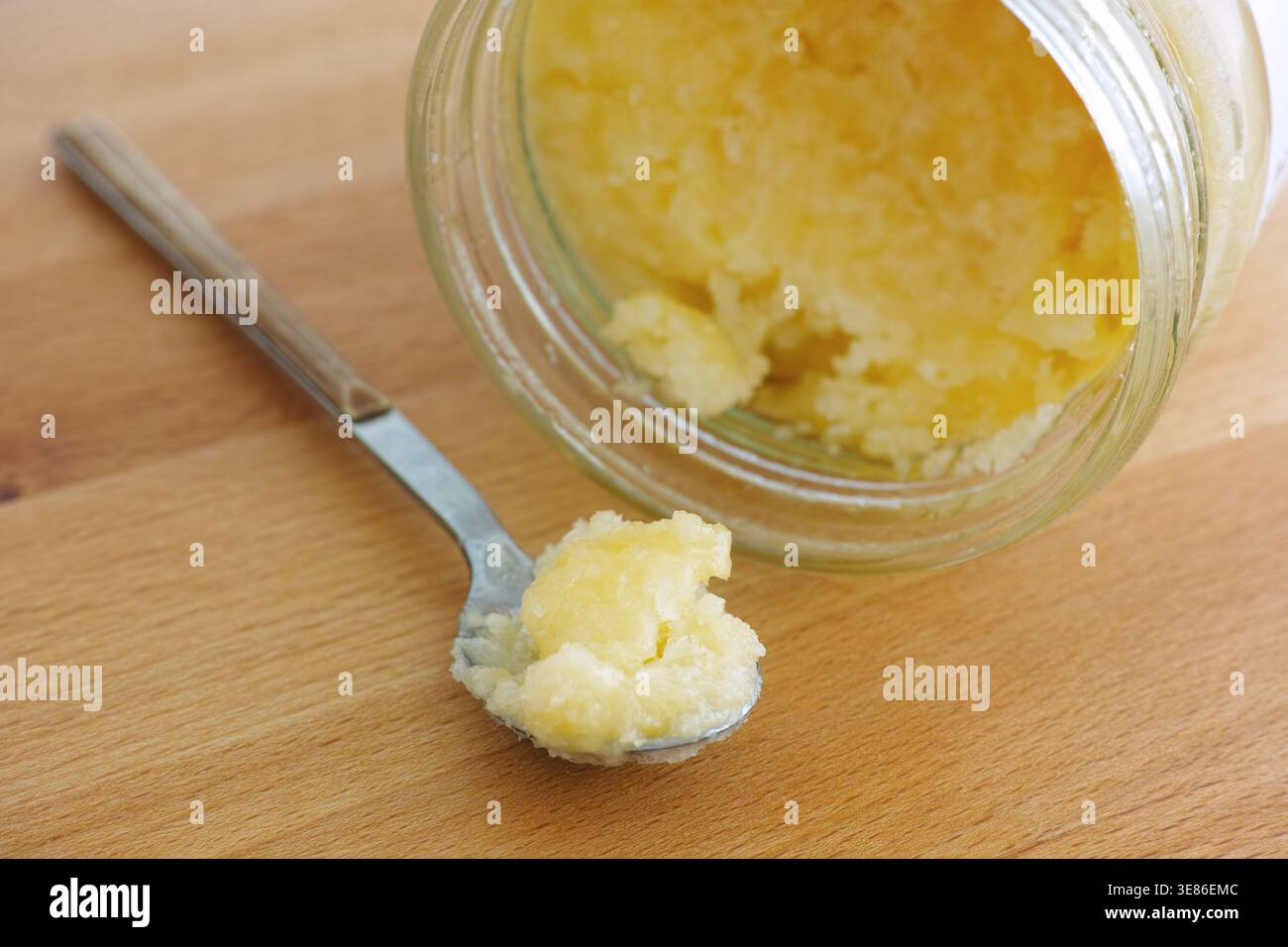 Un piccolo cucchiaio di metallo tiene una porzione di olio di cocco giallo accanto a un vaso di vetro aperto su uno sfondo di legno. Primo piano. Foto Stock
