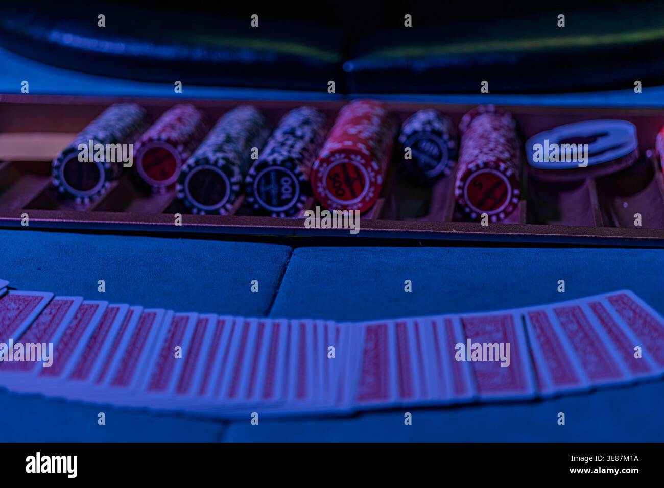 Interno del casinò sotto una vivace luce blu, scatola centrale con chip con gettoni assortiti, pila di carte da gioco nelle vicinanze, prospettiva leggermente superiore Foto Stock