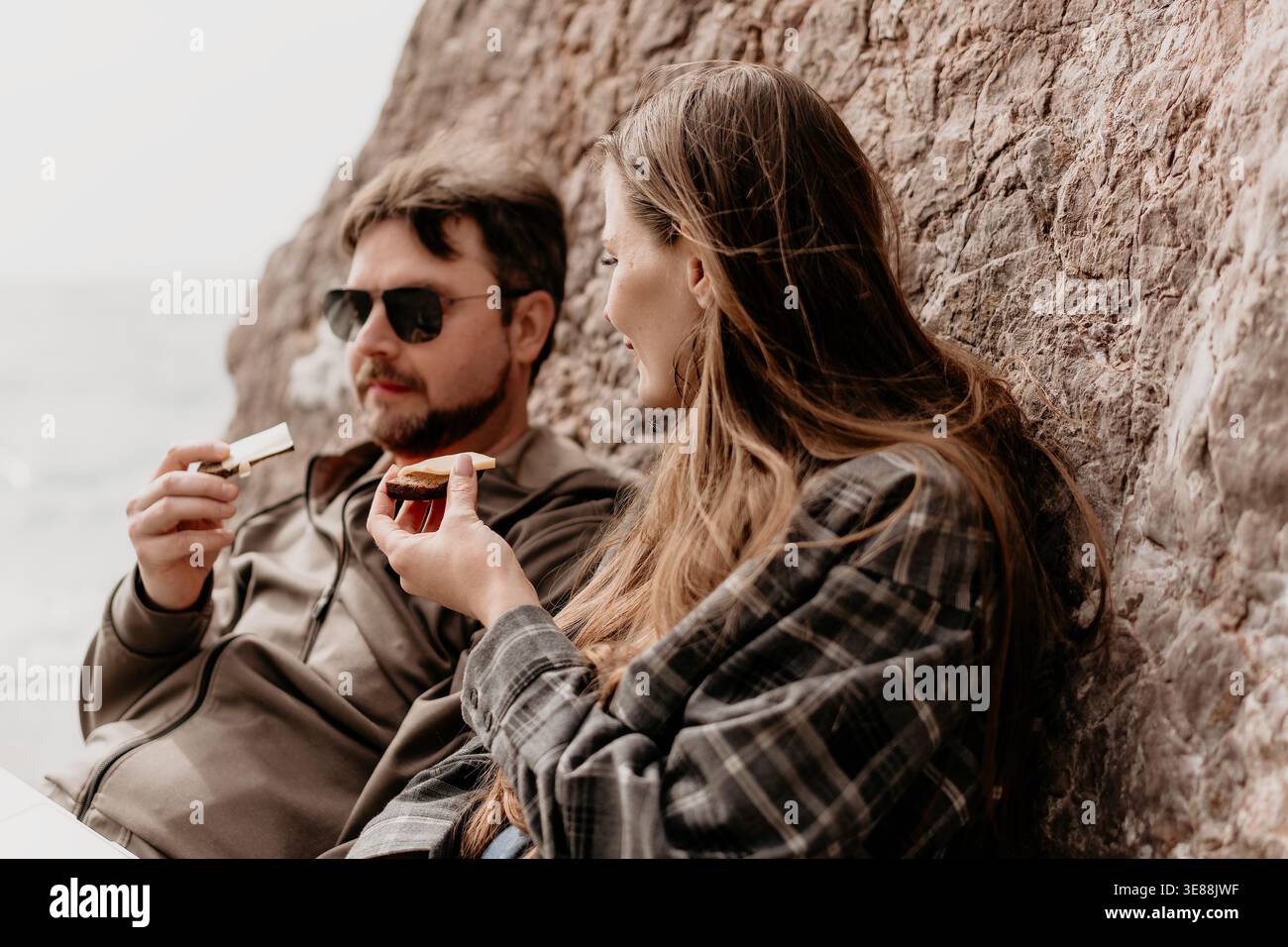 Coppia di spuntini viaggiano in coppia gusta semplici spuntini riposati accanto a una parete di roccia durante il loro informale viaggio all'aperto Foto Stock