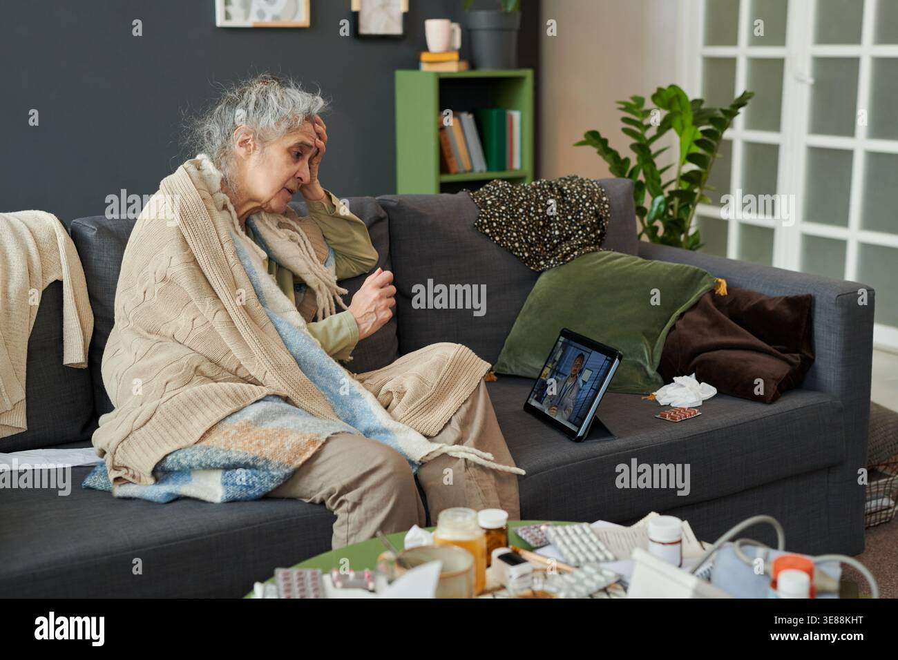 Donna caucasica anziana seduta sul divano, tenendo la testa e consultando il medico su tablet durante l'appuntamento remoto, mostrando malattia e assistenza domiciliare. Utile per telemedicina, marketing senior per l'assistenza sanitaria Foto Stock