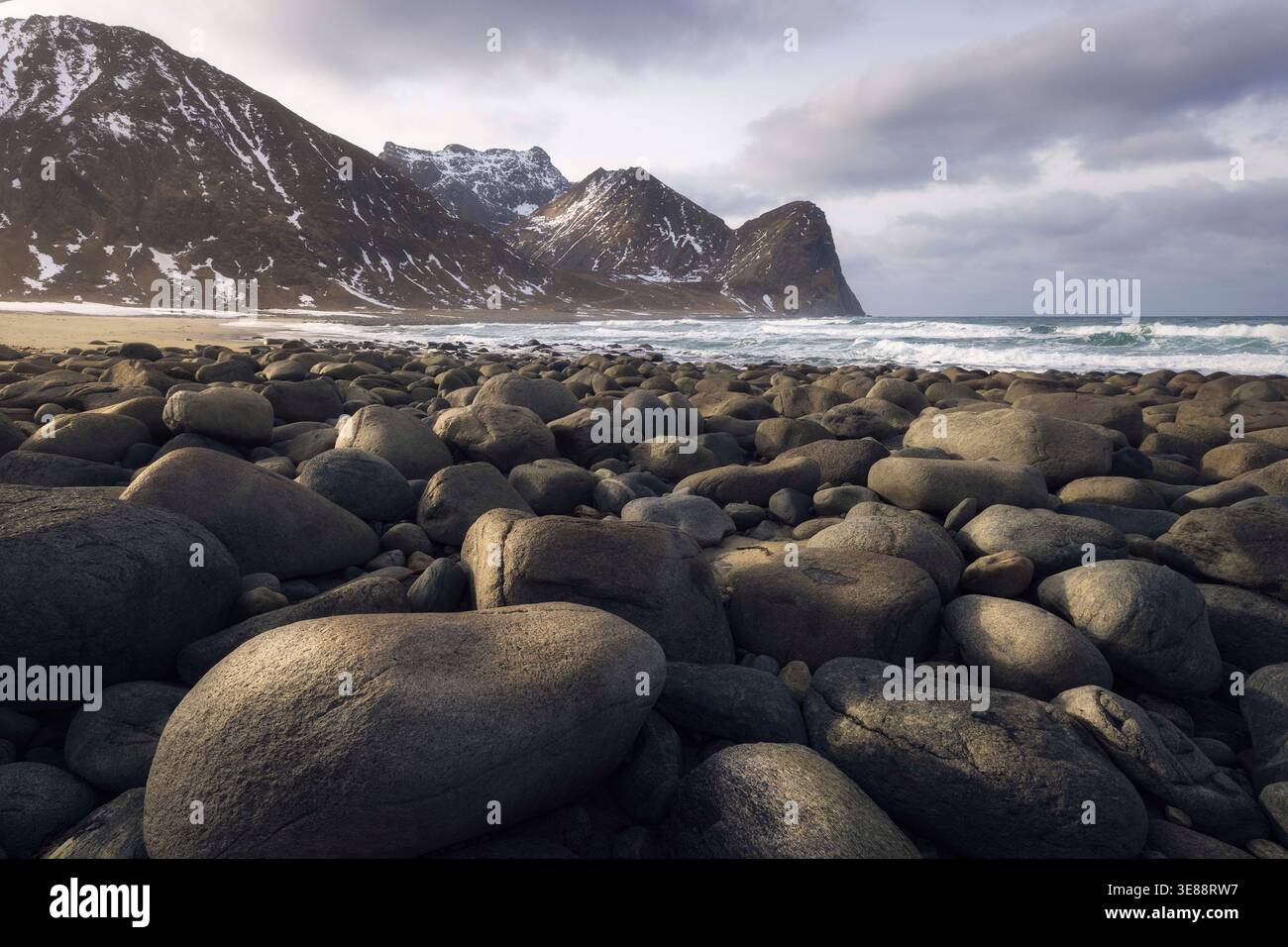 Le onde si infrangono dolcemente sulla costa rocciosa mentre le splendide montagne si innalzano sopra la spiaggia di Unstad. Questo luogo tranquillo rivela la bellezza della natura, creando un tranquillo bancomat Foto Stock