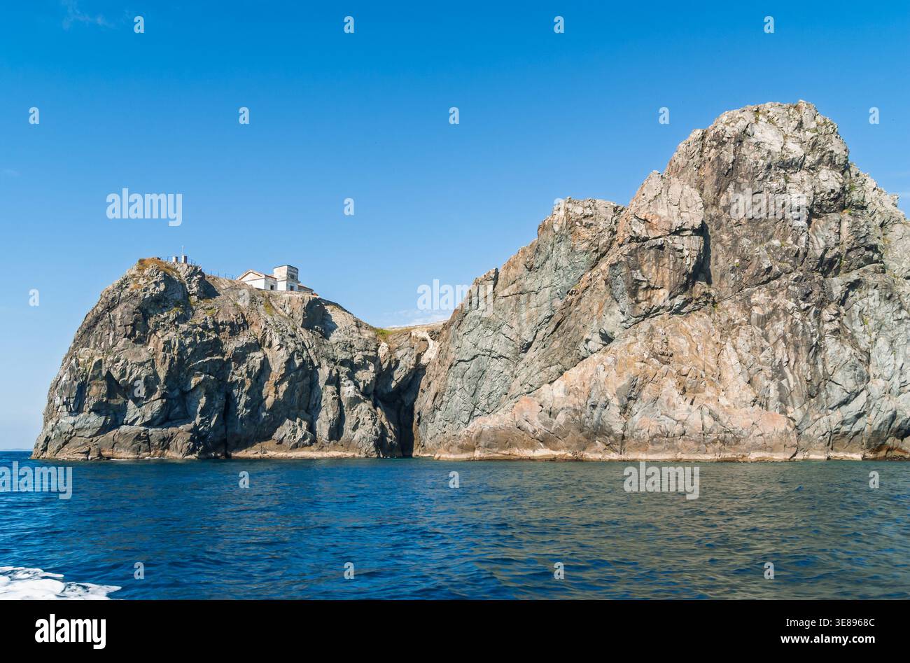 Scogliere costiere caratterizzate da un remoto edificio bianco sulla sua cima, torreggiante sul mare blu profondo in una giornata luminosa Foto Stock