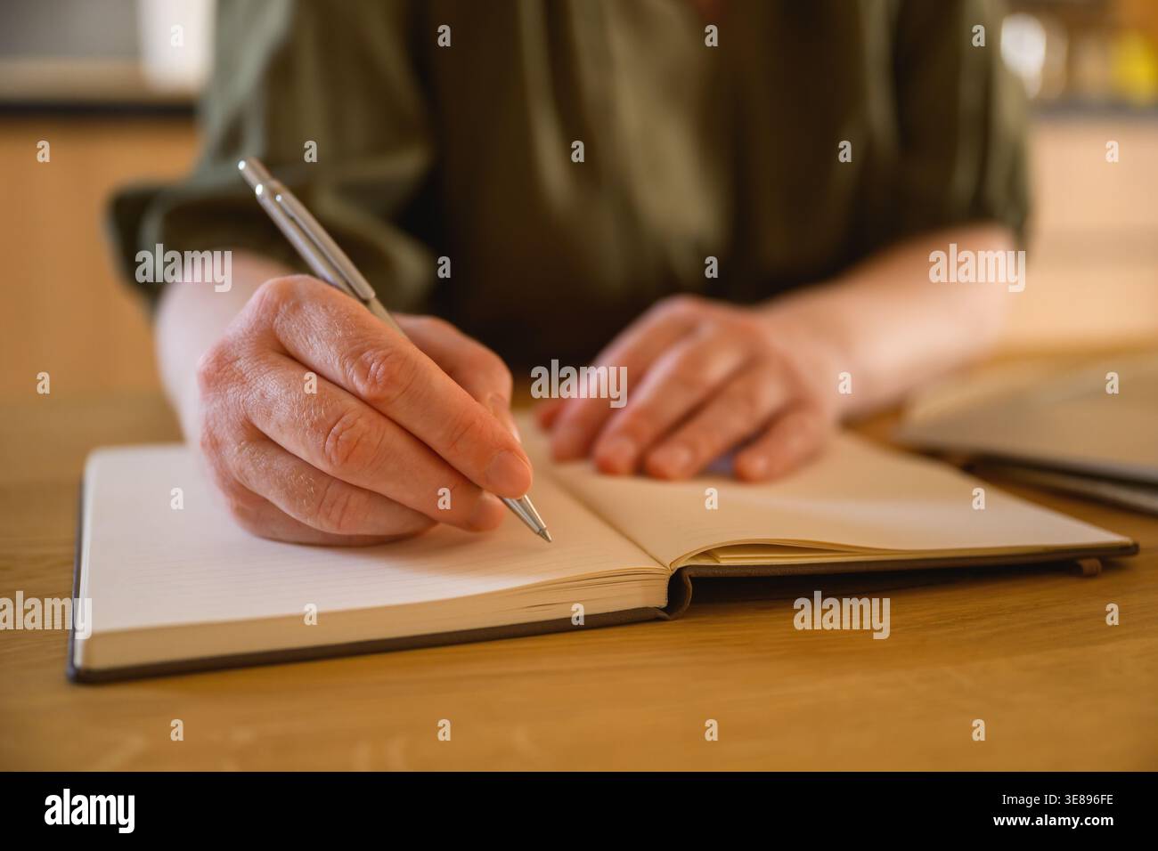 Mani femminili che scrivono su un notebook sulla scrivania in un'accogliente area di lavoro domestica, scrivono, progettano o studiano con un'illuminazione calda e soffusa. Messa a fuoco selettiva. Foto Stock