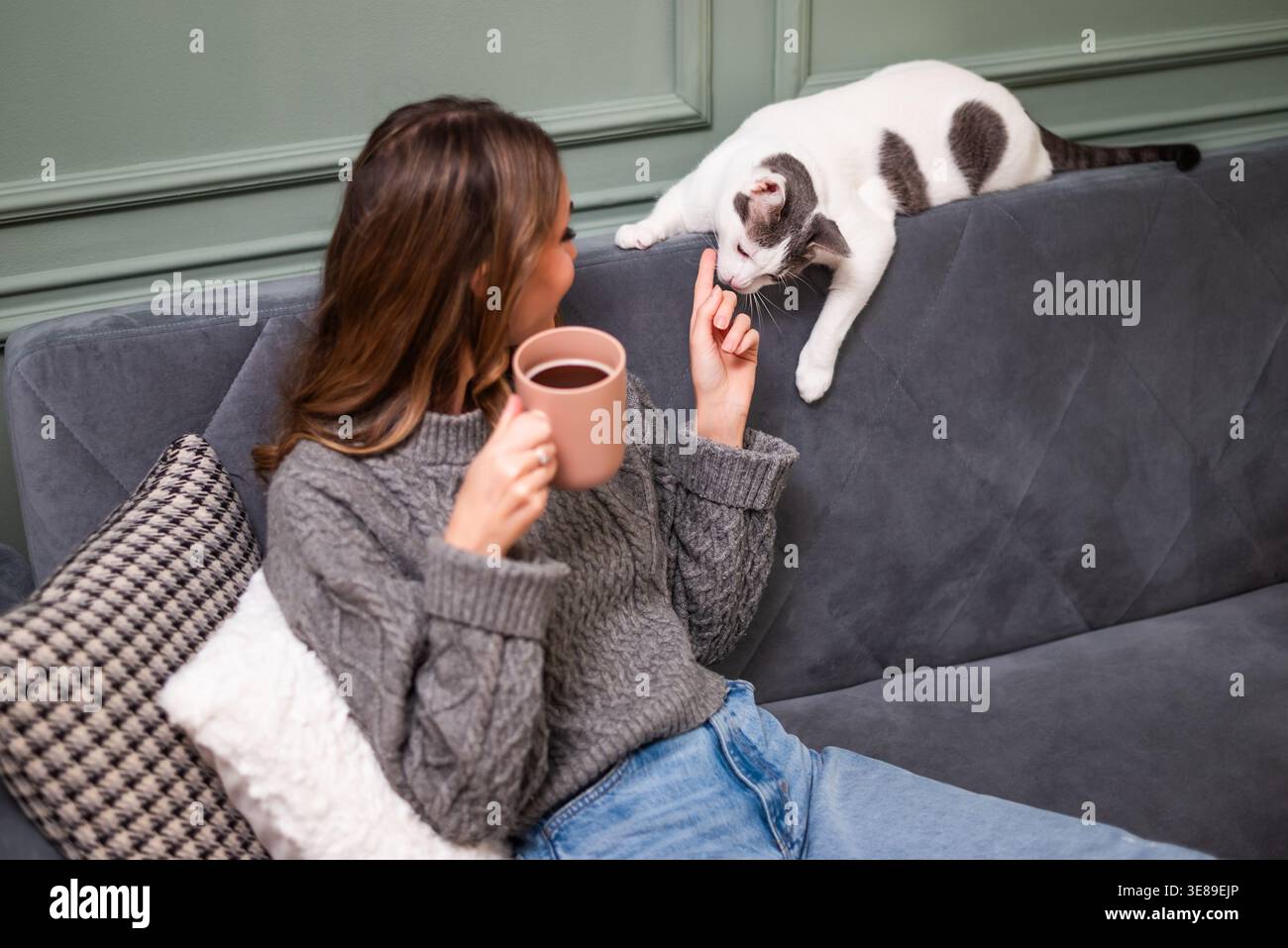 Una donna adulta felice si sta rilassando a casa. È seduta sul divano e beve caffè, mentre gatto è disteso accanto a lei. Foto Stock