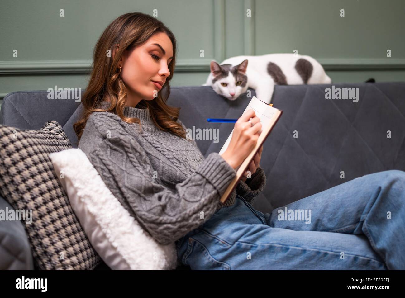 Donna allegra e intima vestita seduto sul divano e scrivendo diario. Il suo gatto è disteso accanto a lei. Foto Stock