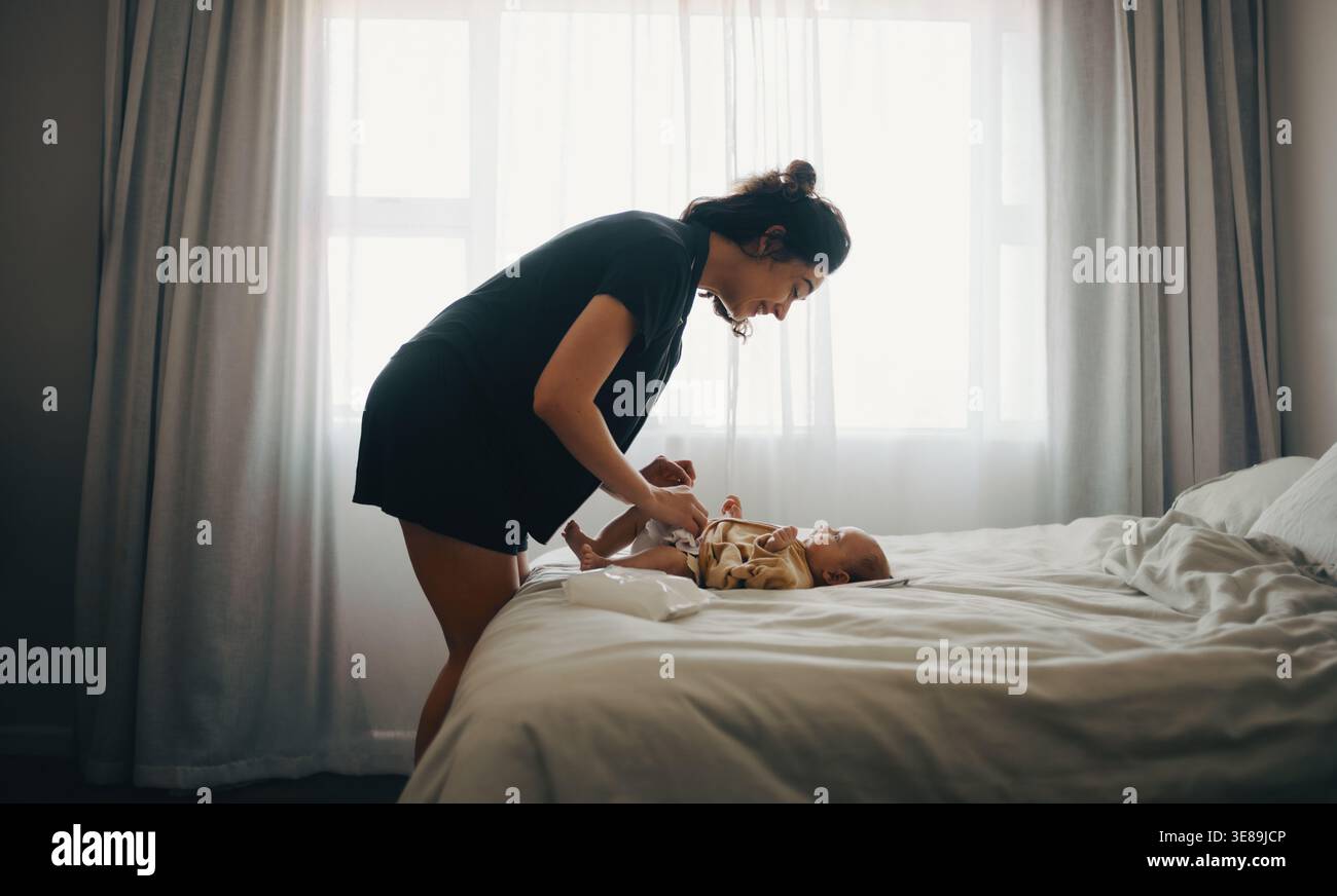 Una madre sorride mentre gioca con il suo bambino su un letto ben fatto in una camera da letto luminosa, catturando un momento caldo e intimo di amore per la famiglia. Foto Stock