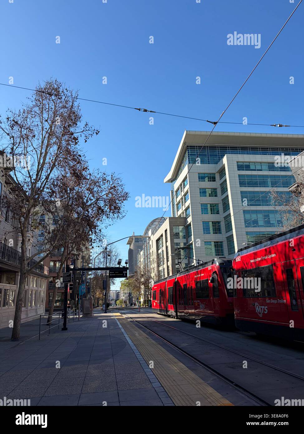Moderno tram rosso MTS che attraversa la strada della città con architettura contemporanea e un cielo azzurro cristallino sullo sfondo a San Diego, California - Immagine stock catturata con smartphone