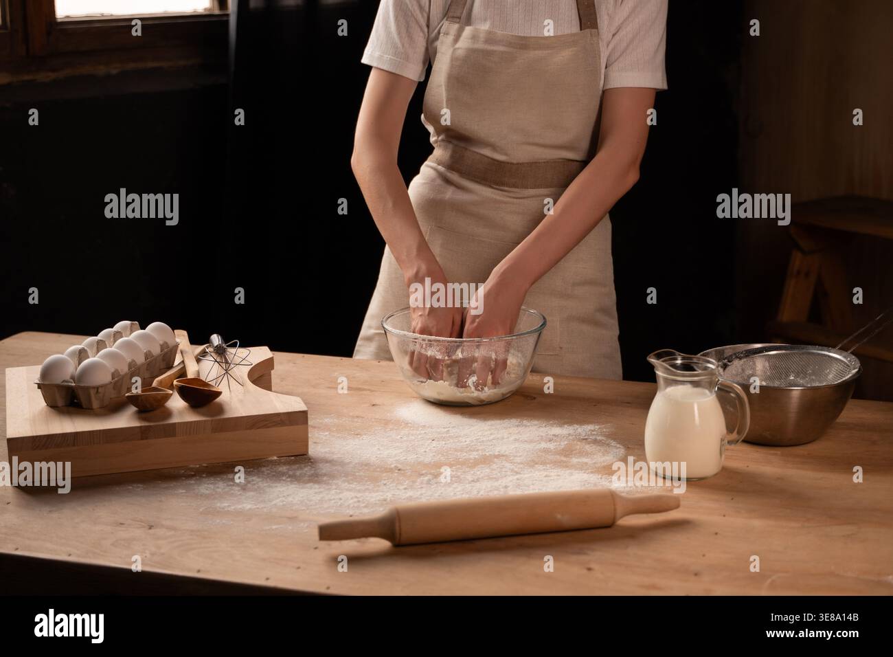 Panettiera che impastava l'impasto in un recipiente di vetro per il pane fatto in casa sul bancone della cucina in legno in stile rustico Foto Stock