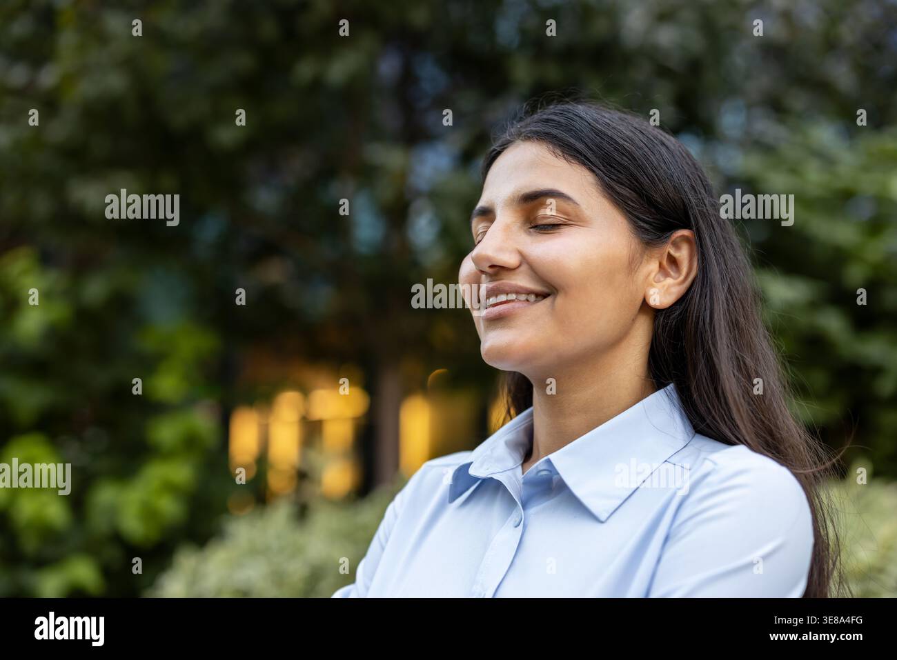 Giovane donna indiana impegnata nella meditazione all'aperto e nella consapevolezza, chiudendo gli occhi felicemente mentre sorride in un tranquillo parco, sentendosi calma ed equilibrata Foto Stock