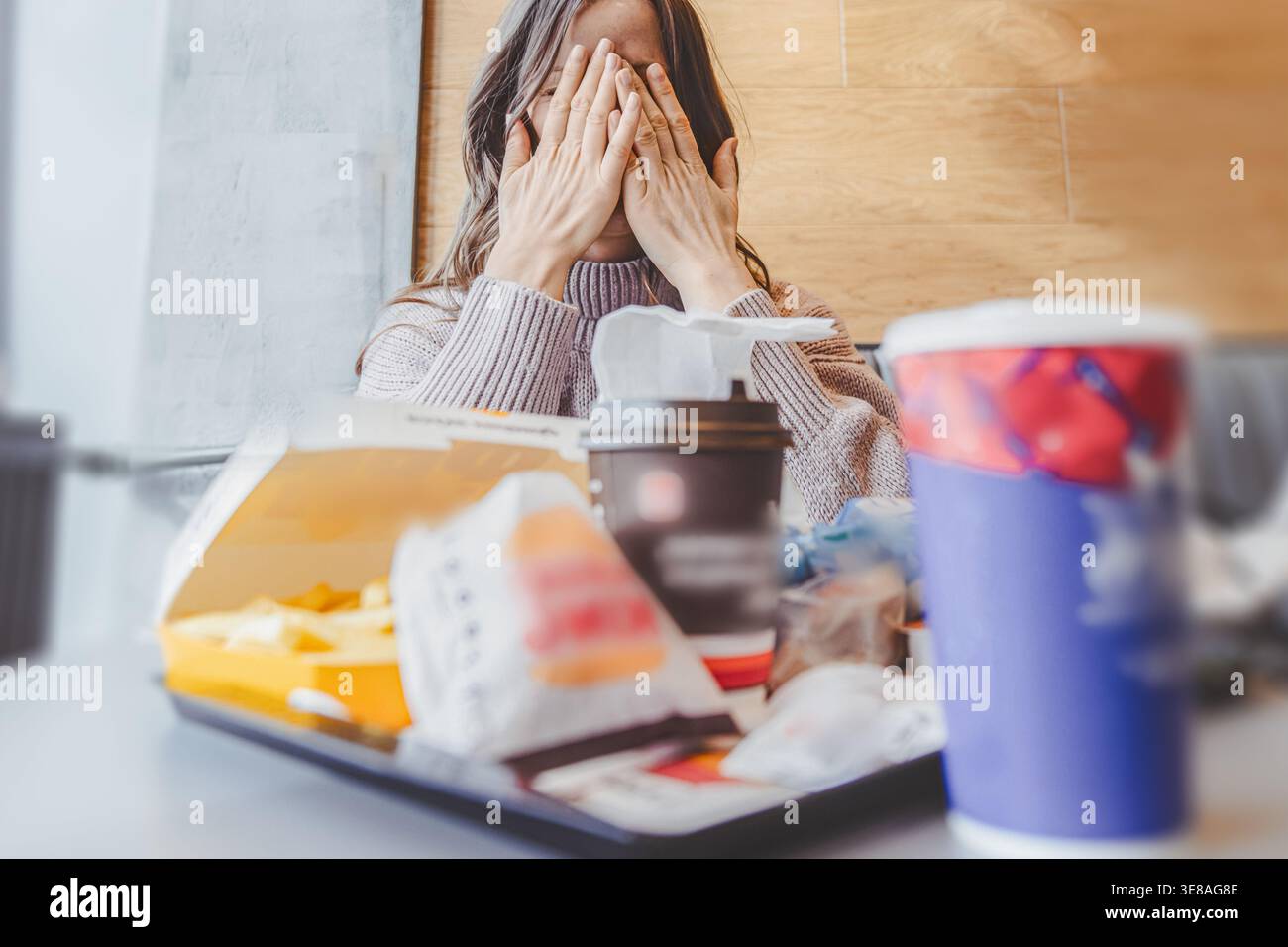 Giovane donna seduta ad un tavolo di fast food con hamburger, patatine fritte e bevande, esitando prima di mangiare. L'immagine evidenzia il processo decisionale e le scelte alimentari Foto Stock