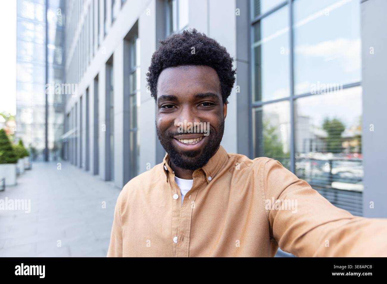 Il giovane imprenditore nero sorridente scatta un selfie sicuro all'aperto in un ambiente urbano moderno, con la testa candida con gli edifici degli uffici sullo sfondo luminoso diurno, autentico e allegro Foto Stock