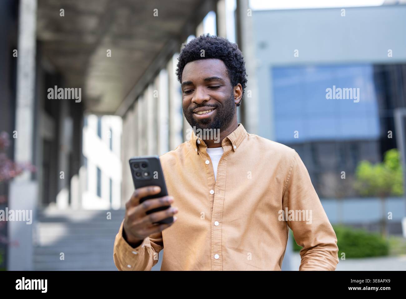 Giovane uomo di discendenza africana sorridente e con in mano un telefono cellulare, coinvolgendo i contenuti digitali mentre si trova in un ambiente urbano durante il giorno Foto Stock