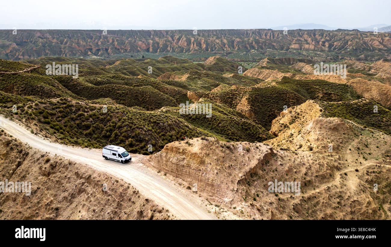 Un camper bianco percorre una strada sterrata lungo un crinale nel Granada Geopark. Questa vista aerea cattura la vastità dei calanchi, delle colline erose, e. Foto Stock