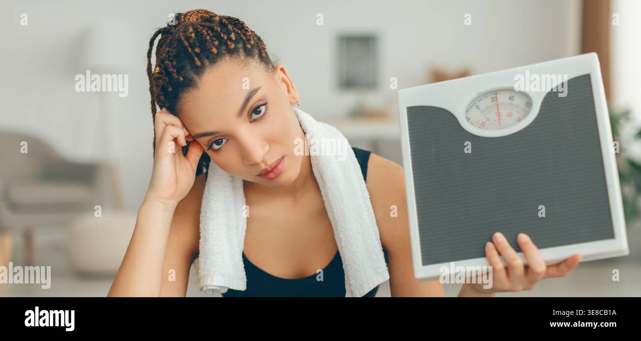 Frustrazione per la bilancia di peso mostrata dalla donna nell'outfit da training che tiene la bilancia in salotto Foto Stock