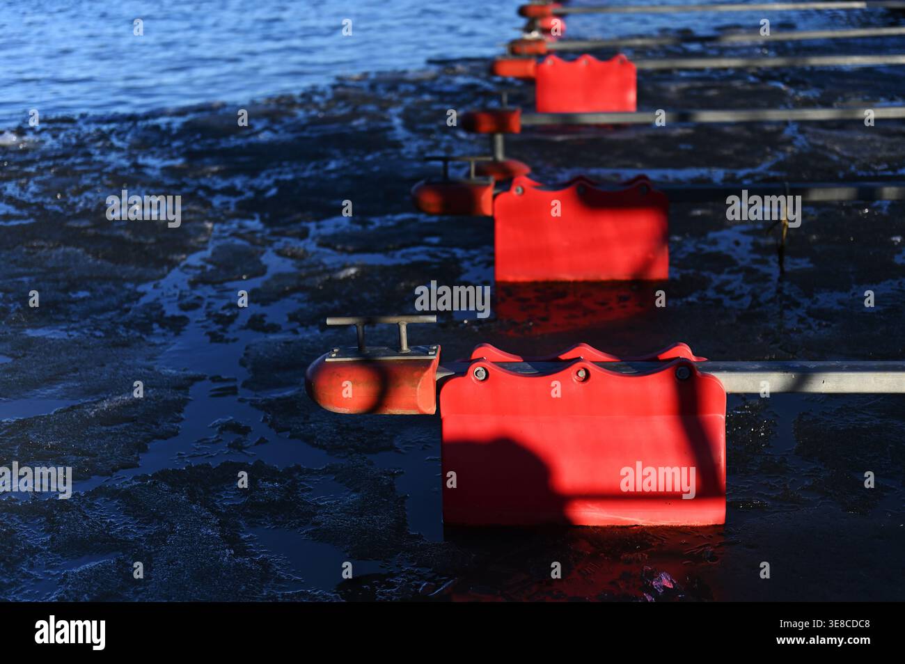 Le barriere rosse della corsia di nuoto si estendono su un lago parzialmente ghiacciato, creando forti linee prospettiche e un contrasto con il ghiaccio blu scuro in un ambiente tranquillo Foto Stock