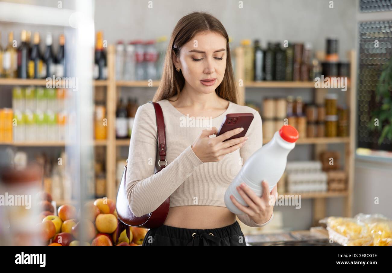 L'acquirente donna verifica l'idoneità - scansiona il codice QR sull'etichetta del flacone di latte nel negozio di alimentari Foto Stock
