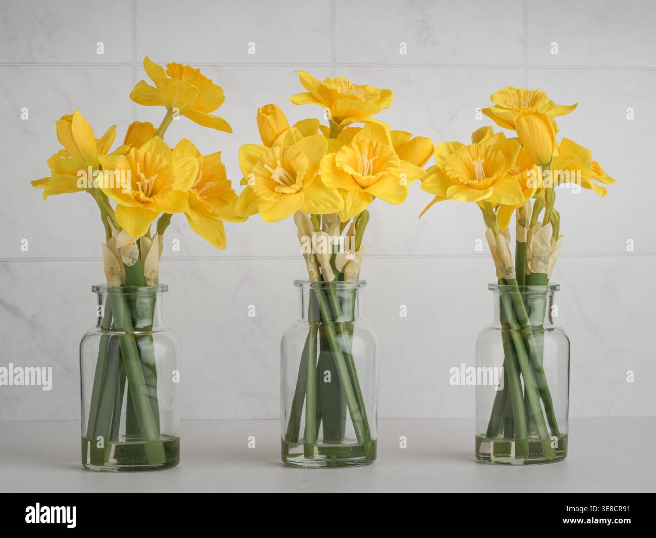 Tre mazzi di allegri narcisi gialli primaverili in vasi di vetro con decorazioni bianche chiare, interni allegri e felici Foto Stock