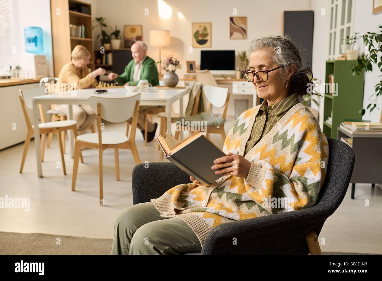 Donna caucasica anziana che legge il libro nella sala di cura mentre la coppia anziana gioca a gioco da tavolo in background, che rappresenta l'invecchiamento attivo e l'uso del marketing della comunità pensionistica Foto Stock